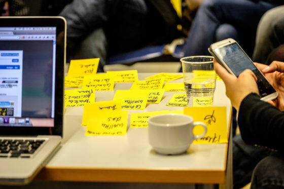 Zu sehen ist ein Tisch mit mehreren gelben Post-it-Zettel darauf, links ein Laptop, rechts eine Hand, die ein Smarphone hält.