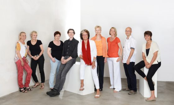 mehrere Menschen stehen in einem weißen Raum, eineige sitzen auf einem weißen Block.