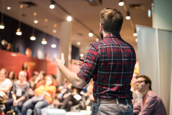 Ein Mann steht vor Publikum und spricht.