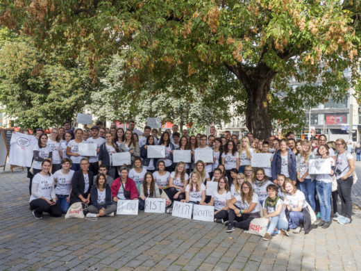 Eine größere Gruppe von Menschen steht und sitzt auf einem Platz unter einem Baum und hält Schilder in die Kamera.