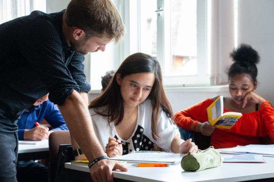 Zwei Schülerinnen sitzen an einem Tisch im Klassenraum. Eine Schülerin liest in einem Buch. Der Lehrer stützt sich auf dem Tisch ab. Schülerin und Lehrer schauen beide auf ein Papier.