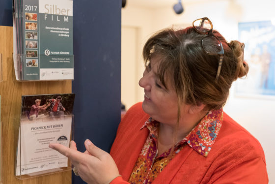 Eine Frau steht vor zwei Halterungen für Broschüren und zeigt mit der linken Hand auf eine der Broschüren.