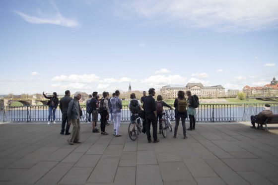 Eine Gruppe von Menschen steht um einen Stadtführer herum am Ufer eines Flusses.
