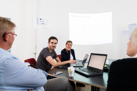 Ein Mann sitzt mit anderen Personen an einem Tisch, vor ihm ein Laptop.