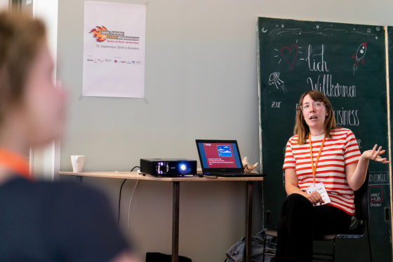Eine Frau im geringelten T-Shirt sitzt neben einem Beamer und erklärt etwas.