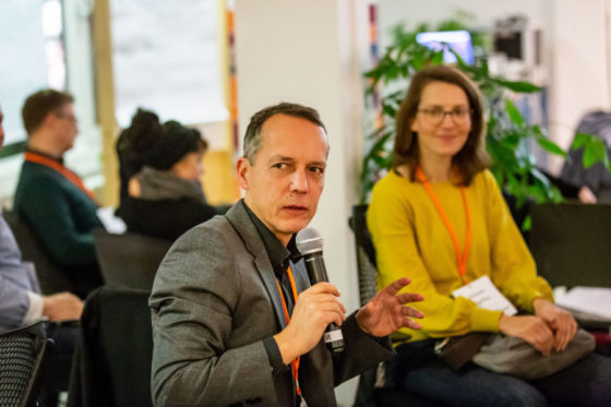 Ein Mann spricht in ein Mikrofon, im Hintergrund sitzt eine Frau.