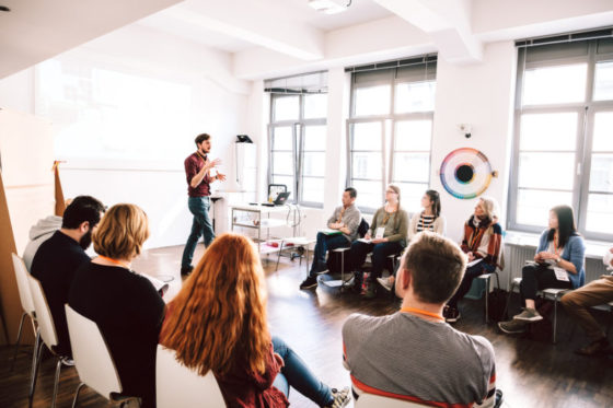 Ein junger Mann steht in einem Stuhlkreis und erklärt etwas.