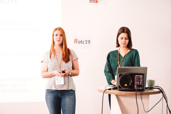 Zwei Frauen stehen vor einer Wand mit dem hashtag #otc19