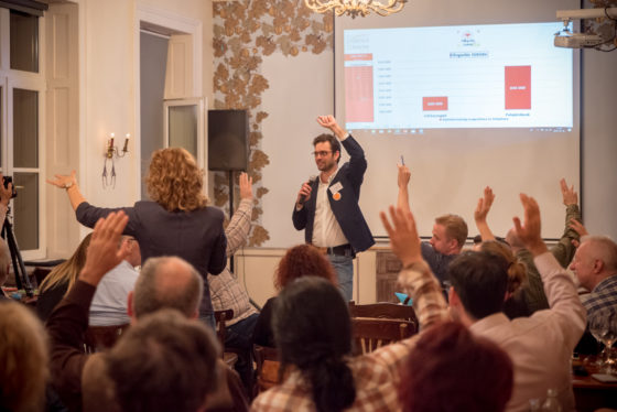 Ein Mann mit Mikrofon steht vor mehreren Menschen, die per Handzeichen abstimmen.