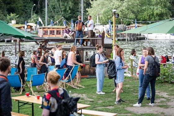 Menschen stehen auf einer Wiese, im Hintergrund ein Holzfloß