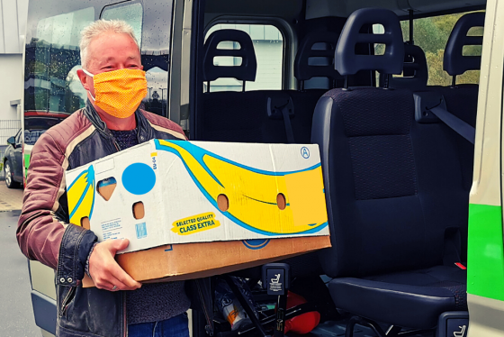 Auf dem Foto ist ein älterer Mann mit einer Maske zu sehen. Er steht vor einem Transportbus und hält einen Bananen-Karton in den Händen.